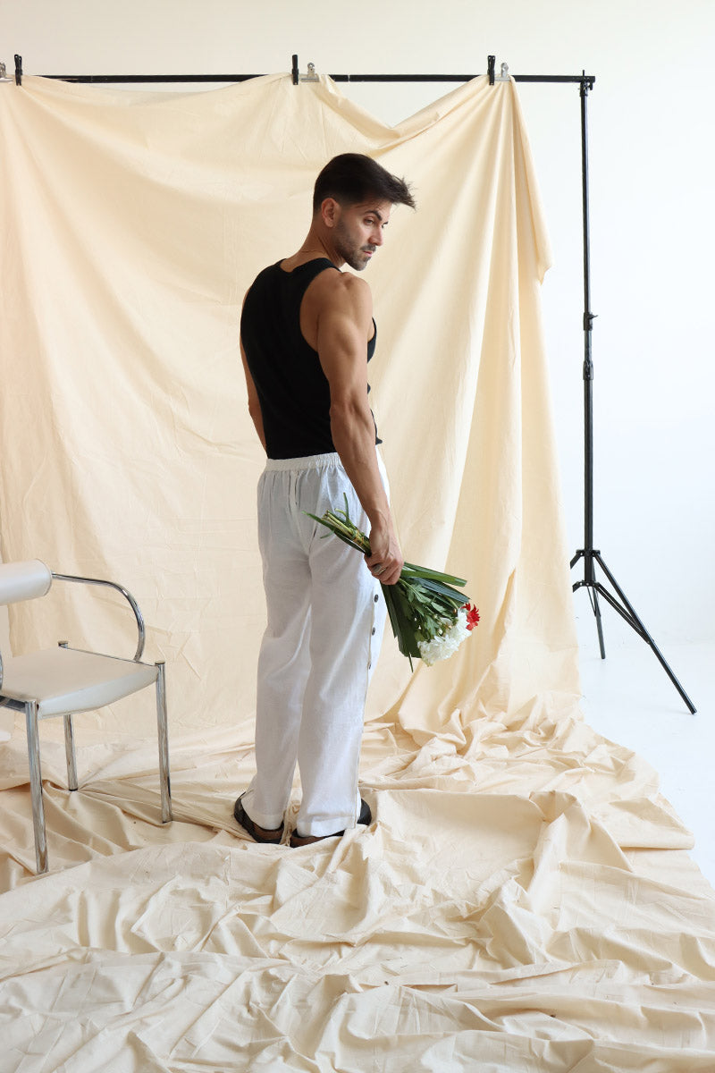 A person standing in front of a neutral backdrop wearing white linen button-up pants with a flat front and elasticated waist. The person has their hands in their pockets, and the image showcases a side slit detail with a line of buttons on the pants.
