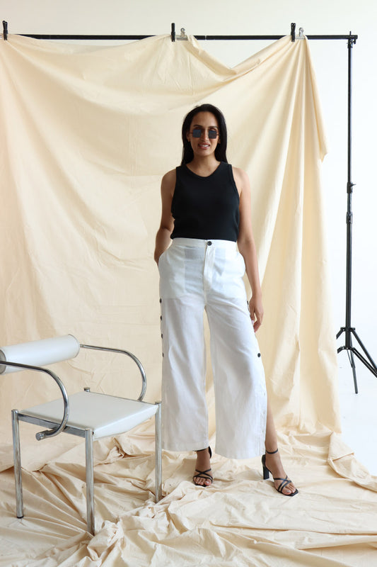 A person standing in front of a neutral backdrop wearing white linen button-up pants with a flat front and elasticated waist. The person has their hands in their pockets, and the image showcases a side slit detail with a line of buttons on the pants.