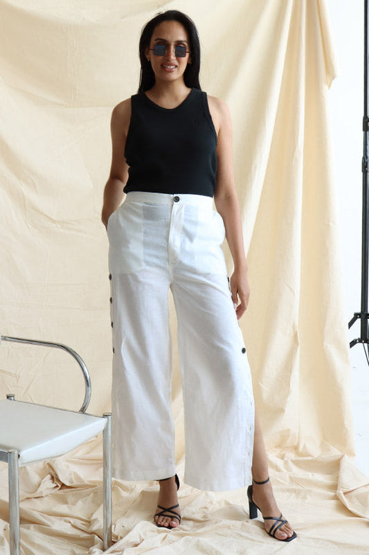 A person standing in front of a neutral backdrop wearing white linen button-up pants with a flat front and elasticated waist. The person has their hands in their pockets, and the image showcases a side slit detail with a line of buttons on the pants.