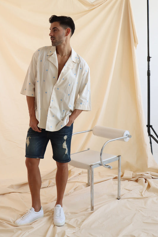 A man wearing a cream cotton shirt with embroidered flowers and a camp collar, paired with blue denim shorts.