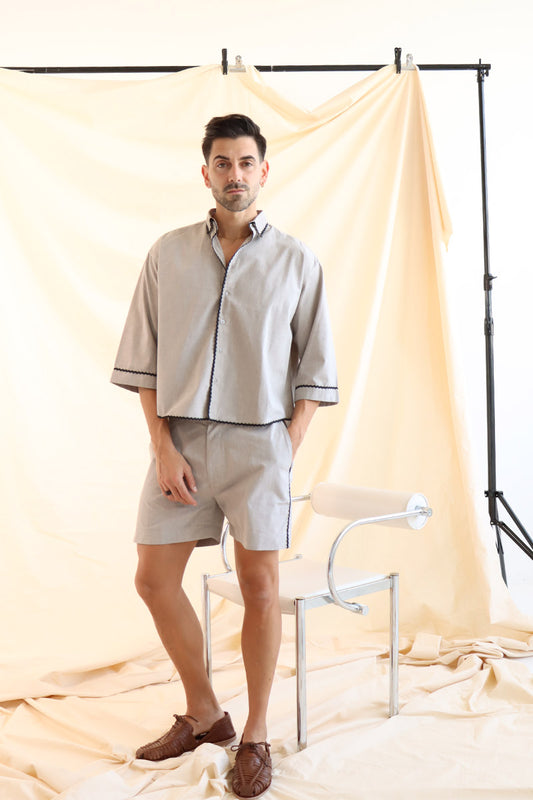 A male model wearing a beige linen short set with a cropped shirt and short shorts, standing in front of a neutral backdrop.