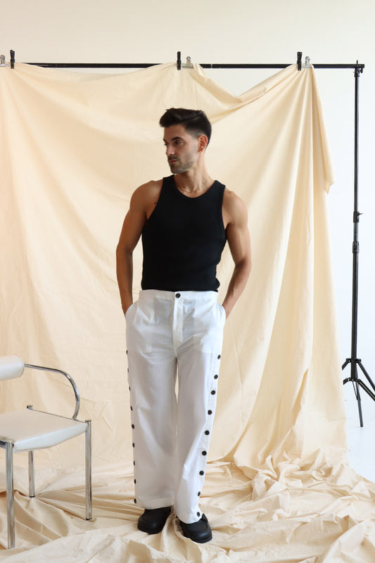 A person standing in front of a neutral backdrop wearing white linen button-up pants with a flat front and elasticated waist. The person has their hands in their pockets, and the image showcases a side slit detail with a line of buttons on the pants.