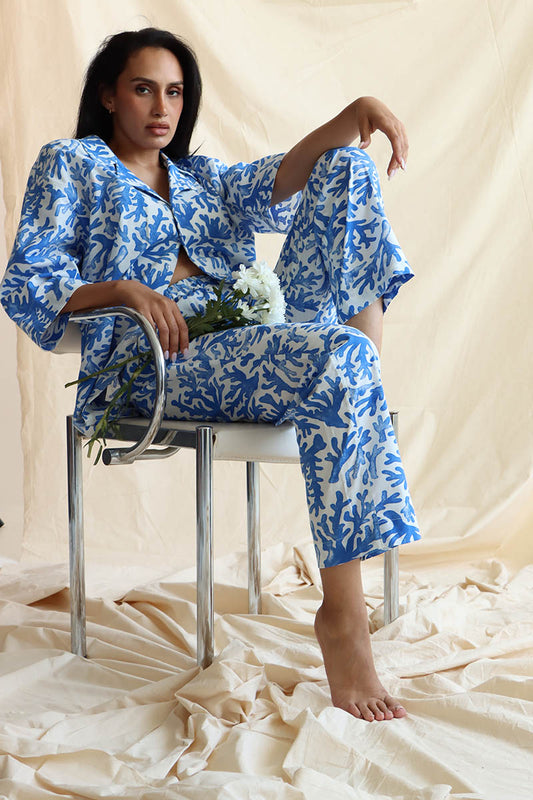 A person seated wearing a coral print linen set with a button-down shirt and 3/4 sleeves, paired with matching relaxed pants with an elastic waistband and drawstring.