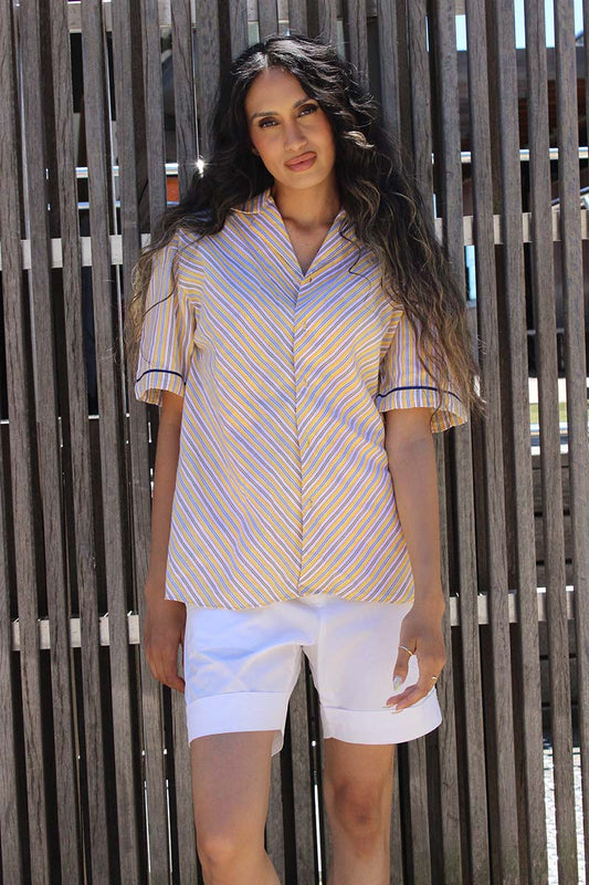 A person wearing a yellow, navy, and white striped short-sleeved shirt with a revere collar and front button closure, paired with white shorts and yellow sunglasses, standing in front of a wooden fence.