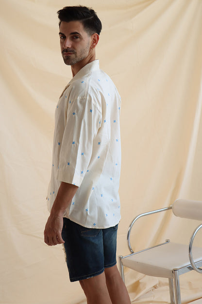 A man wearing a cream cotton shirt with embroidered flowers and a camp collar, paired with blue denim shorts.