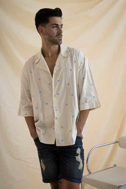 A man wearing a cream cotton shirt with embroidered flowers and a camp collar, paired with blue denim shorts.