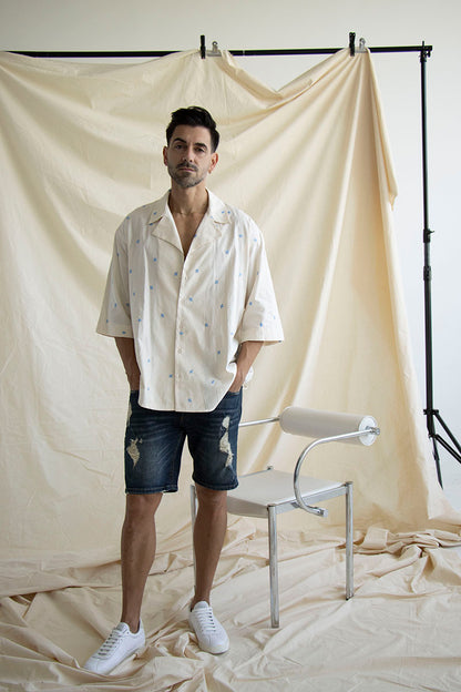 A person wearing a cream cotton shirt with embroidered flowers, short sleeves, and a camp collar. The shirt is displayed in a casual setting with a neutral background.