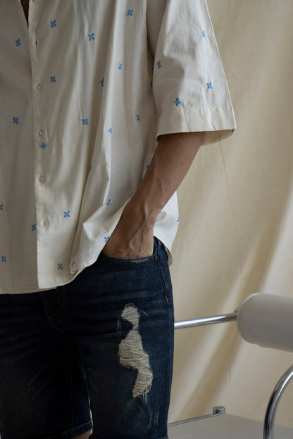 A man wearing a cream cotton shirt with embroidered flowers and a camp collar, paired with blue denim shorts.