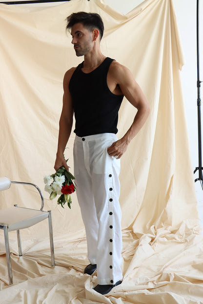 A person standing in front of a neutral backdrop wearing white linen button-up pants with a flat front and elasticated waist. The person has their hands in their pockets, and the image showcases a side slit detail with a line of buttons on the pants.