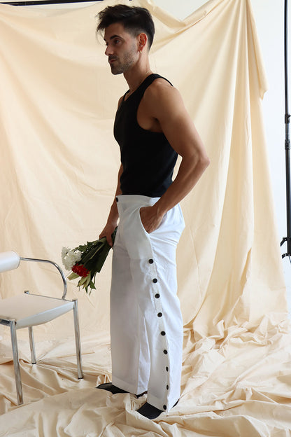 A person standing in front of a neutral backdrop wearing white linen button-up pants with a flat front and elasticated waist. The person has their hands in their pockets, and the image showcases a side slit detail with a line of buttons on the pants.