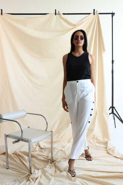 A person standing in front of a neutral backdrop wearing white linen button-up pants with a flat front and elasticated waist. The person has their hands in their pockets, and the image showcases a side slit detail with a line of buttons on the pants.