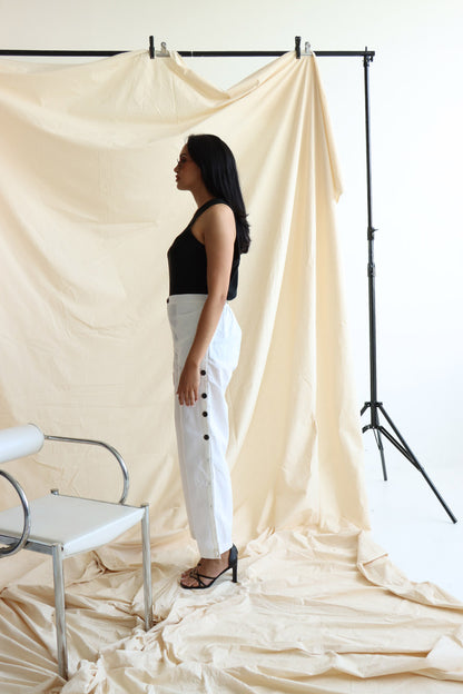 A person standing in front of a neutral backdrop wearing white linen button-up pants with a flat front and elasticated waist. The person has their hands in their pockets, and the image showcases a side slit detail with a line of buttons on the pants.