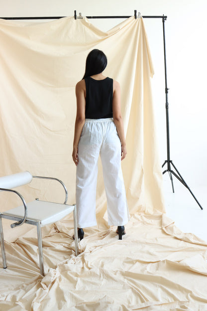 A person standing in front of a neutral backdrop wearing white linen button-up pants with a flat front and elasticated waist. The person has their hands in their pockets, and the image showcases a side slit detail with a line of buttons on the pants.