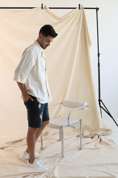 A man wearing a cream cotton shirt with embroidered flowers and a camp collar, paired with blue denim shorts.