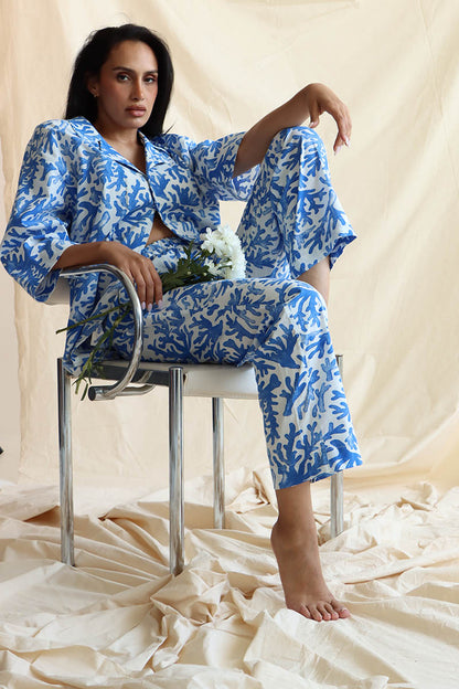 A person seated wearing a coral print linen set with a button-down shirt and 3/4 sleeves, paired with matching relaxed pants with an elastic waistband and drawstring.