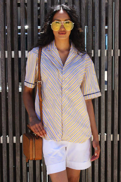 A person wearing a yellow, navy, and white striped short-sleeved shirt with a revere collar and front button closure, paired with white shorts and yellow sunglasses, standing in front of a wooden fence.