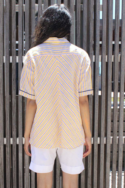 A person wearing a yellow, navy, and white striped short-sleeved shirt with a revere collar and front button closure, paired with white shorts and yellow sunglasses, standing in front of a wooden fence.